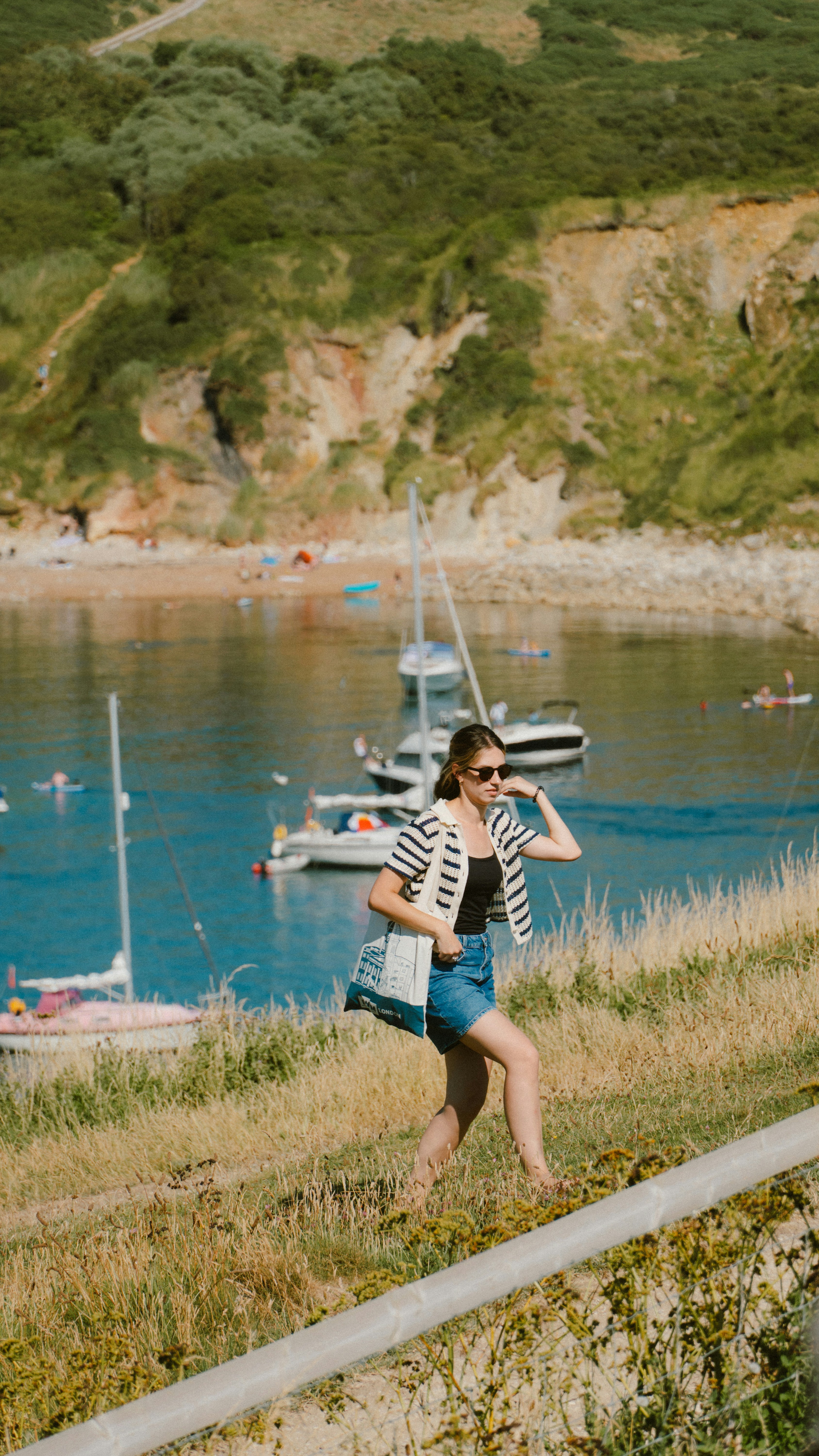 Woman walks up grassy hill overlooking bay with boats.