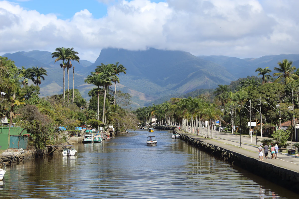 A canal in Paraty lined with palms and moored boats, with green mountains behind