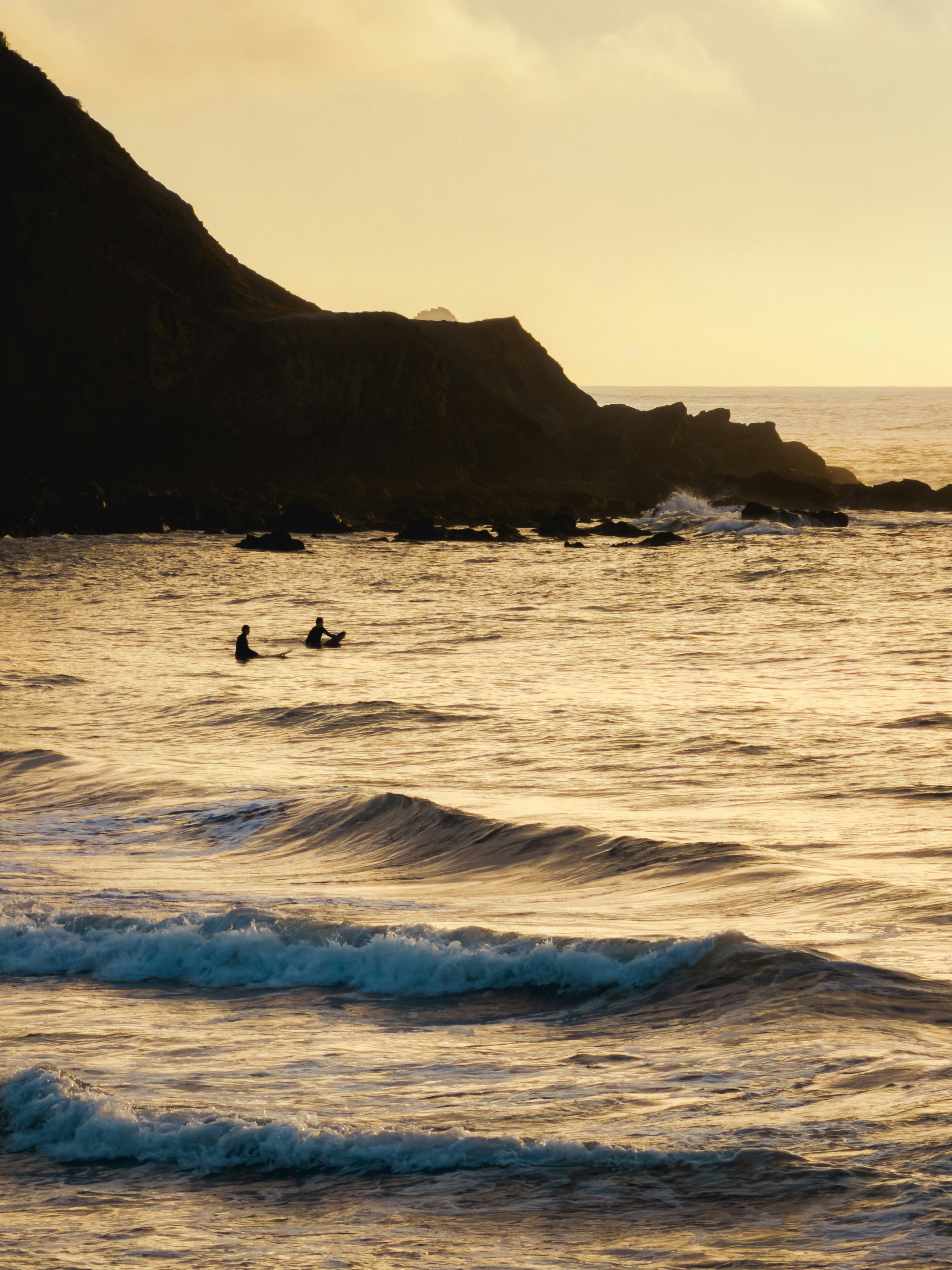 Two surfers wait for waves near a rocky coastline.
