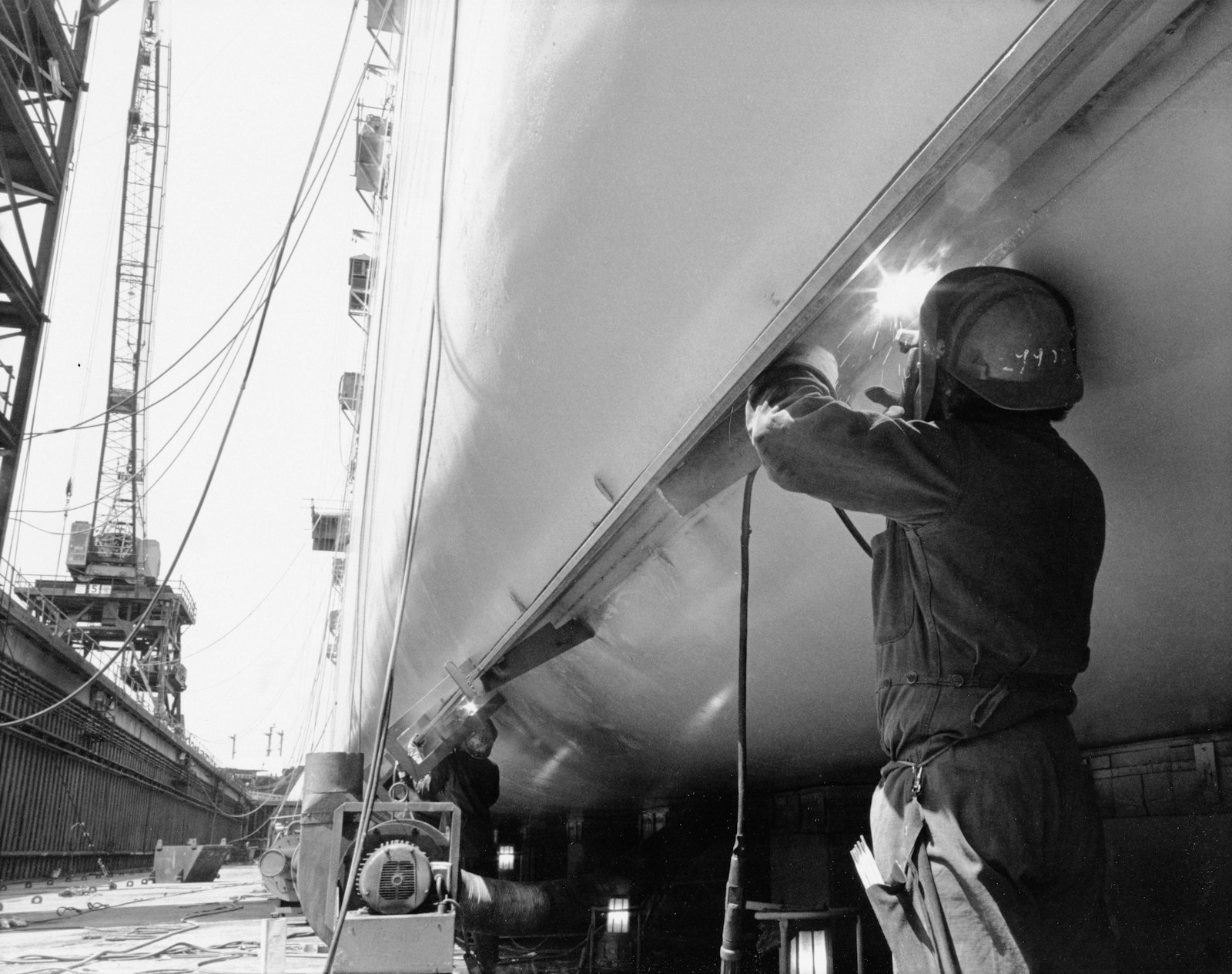 Welder working on a large ship hull in dry dock, the environment where steel plate, restricted ventilation and full PPE stack heat load on top of ambient temperature