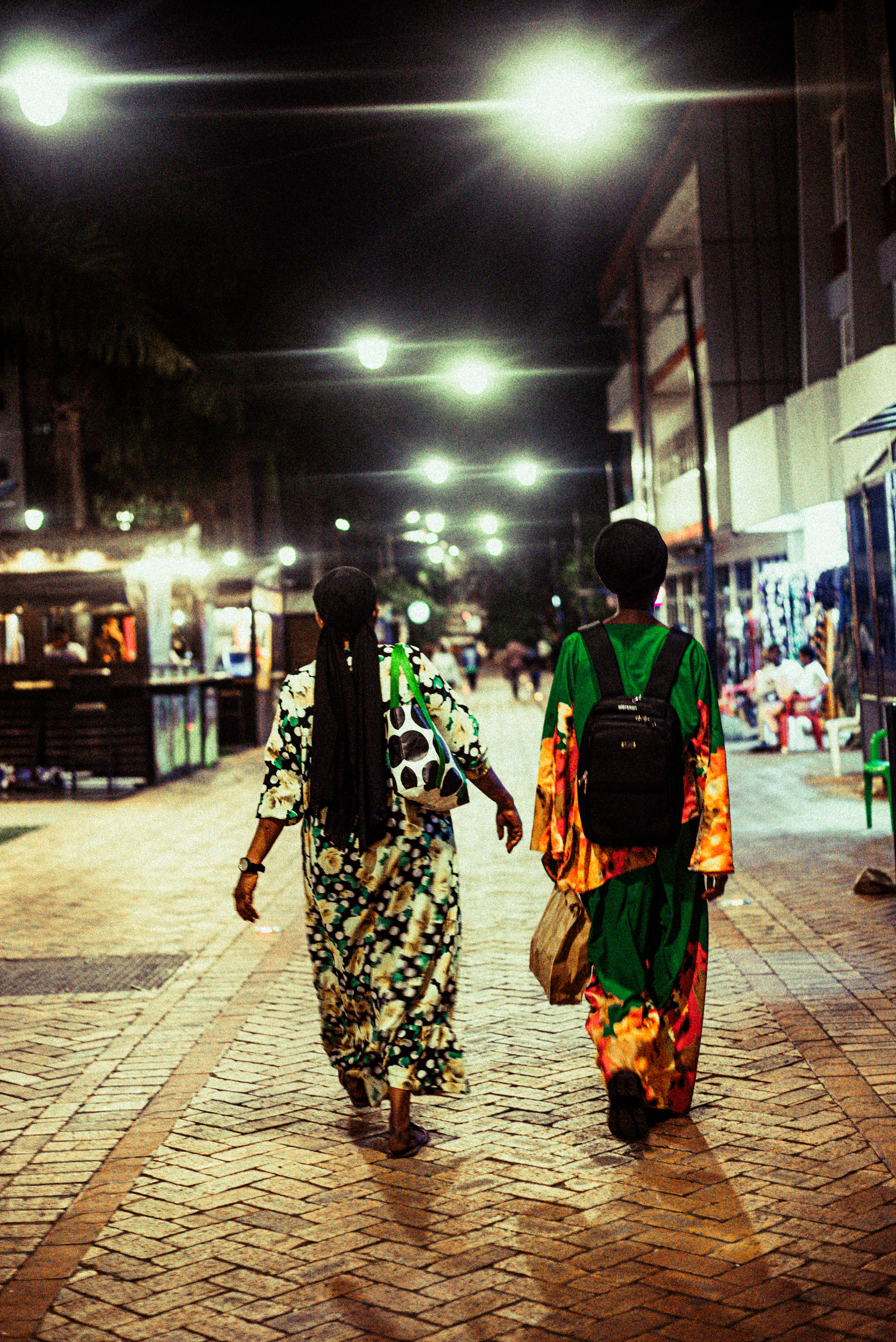 Two women in brightly patterned dresses walking away down a brick-paved pedestrian street in Kigali at night, under strings of street lamps beside lit shopfronts