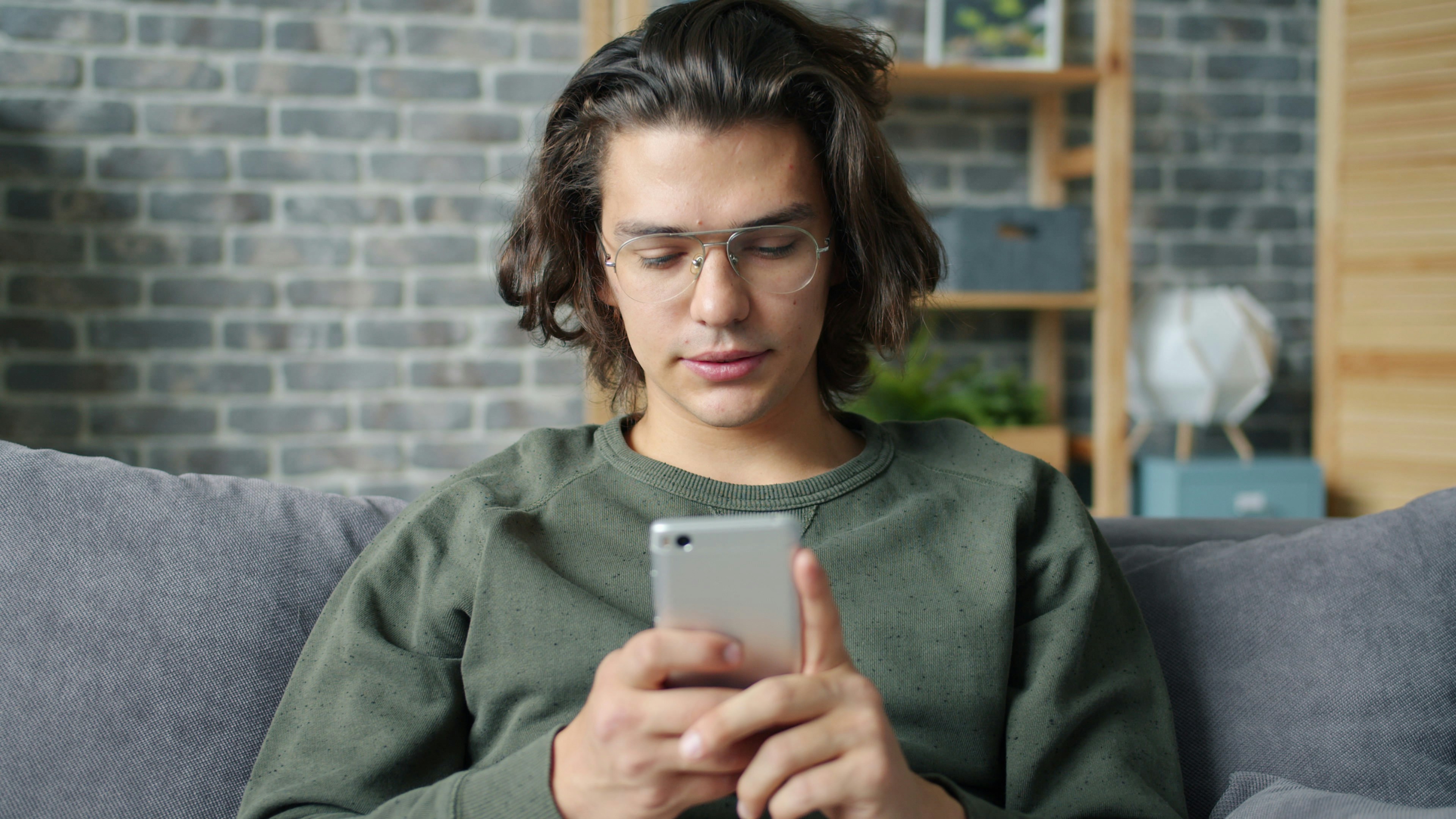 Joven con gafas usando un teléfono inteligente en el sofá.