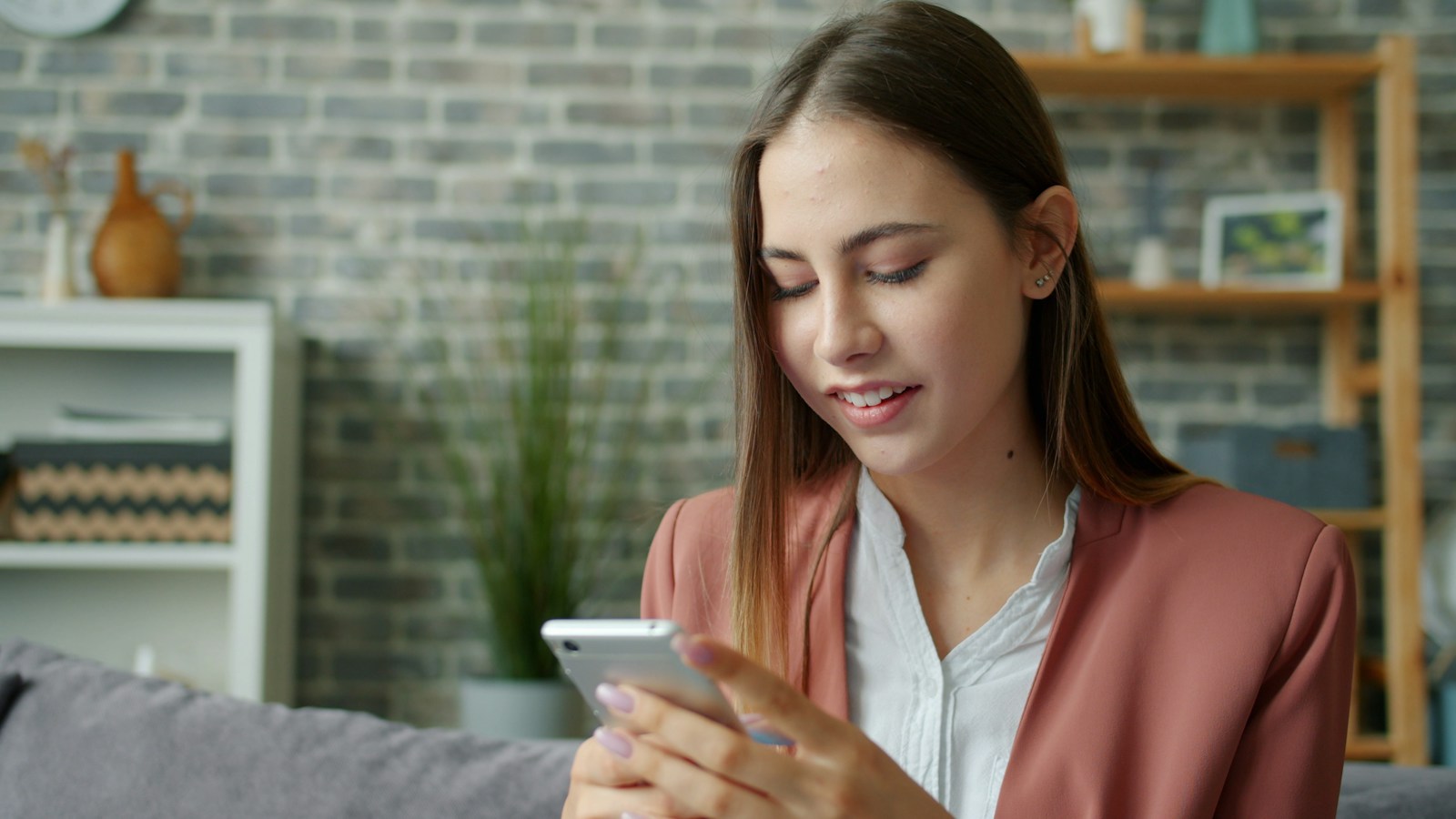 A young woman in a pink blazer sits on a couch in a living room smiling as she looks down at the smartphone she is holding in one hand