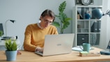 Man in yellow sweater working on laptop at desk.