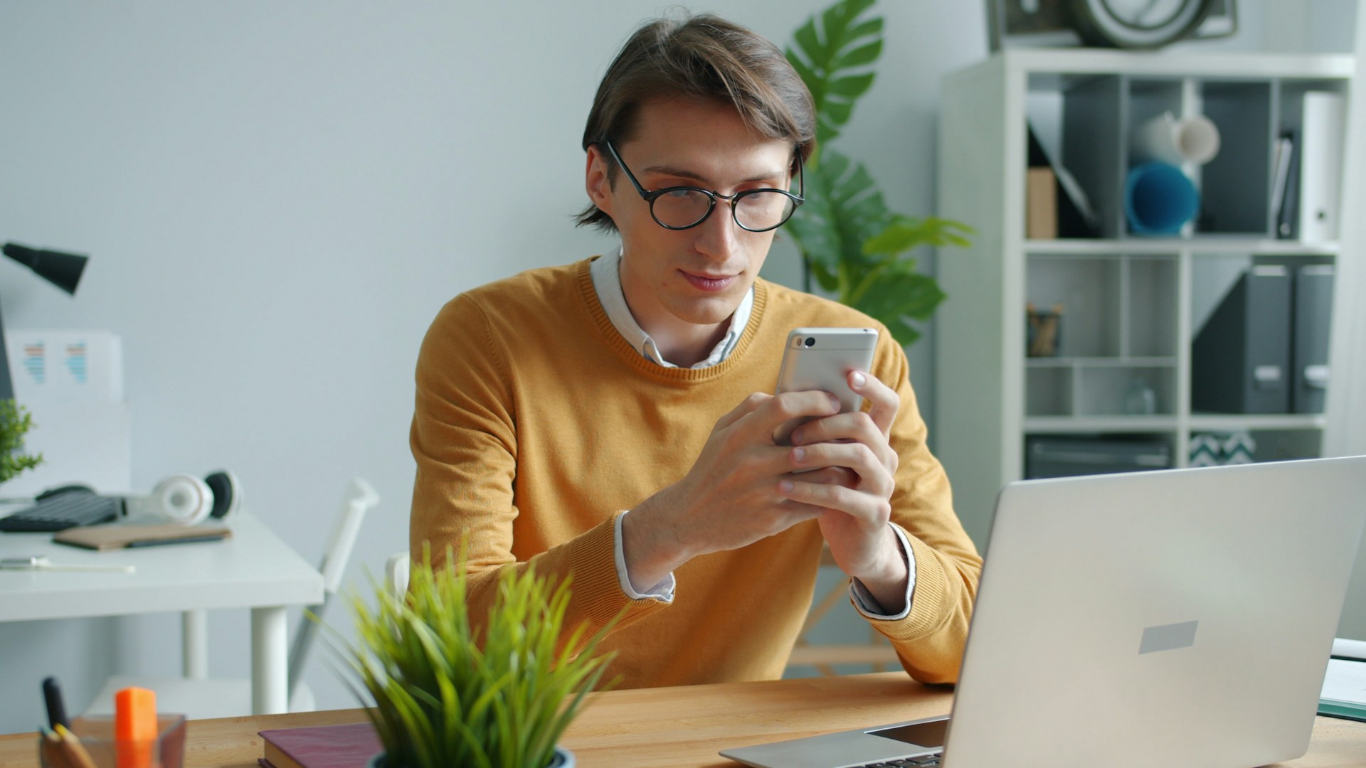 Man in glasses using phone at desk with laptop.
