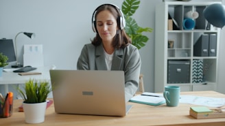 Woman wearing headphones works on laptop at desk.