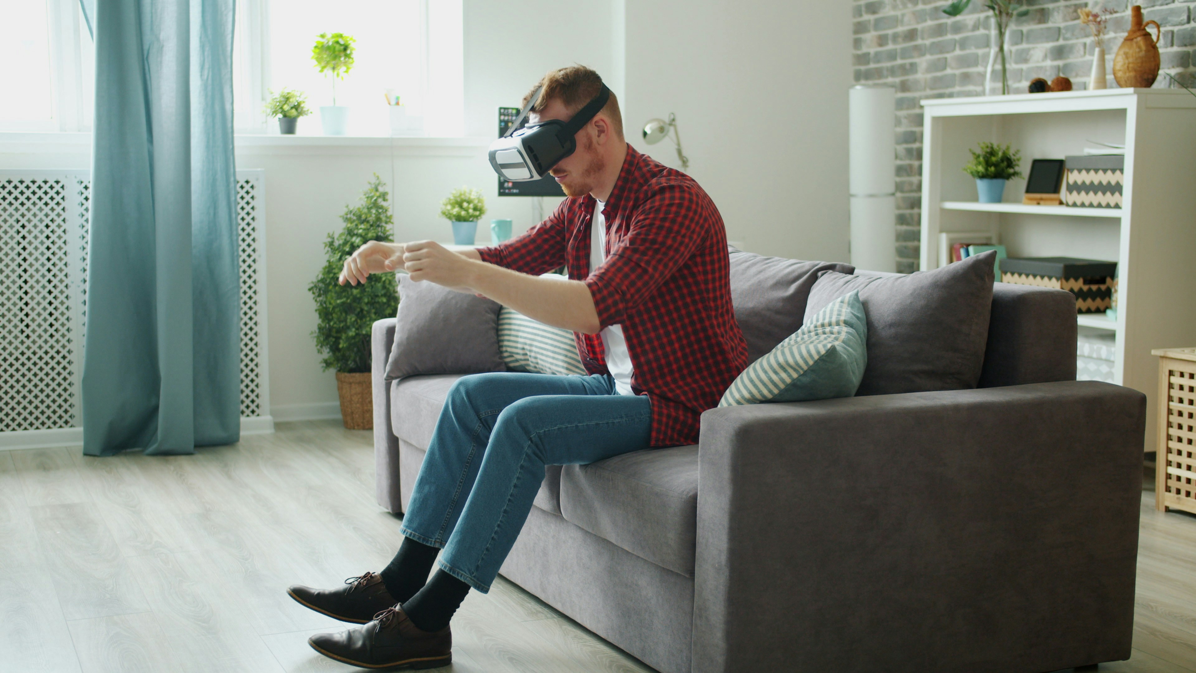 Adult male in casual clothing is enjoying driving activity in virtual reality glasses gesturing sitting on sofa at home. Entertainment and lifestyle concept.