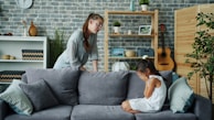 Mother scolds crying daughter on sofa in living room.
