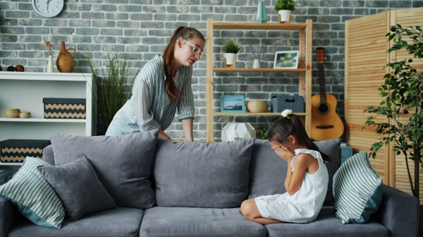 Mother scolds crying daughter on sofa in living room.