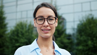 A woman with glasses smiles in front of a building.