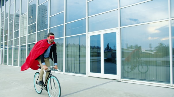 Man in superhero costume rides bicycle past modern building