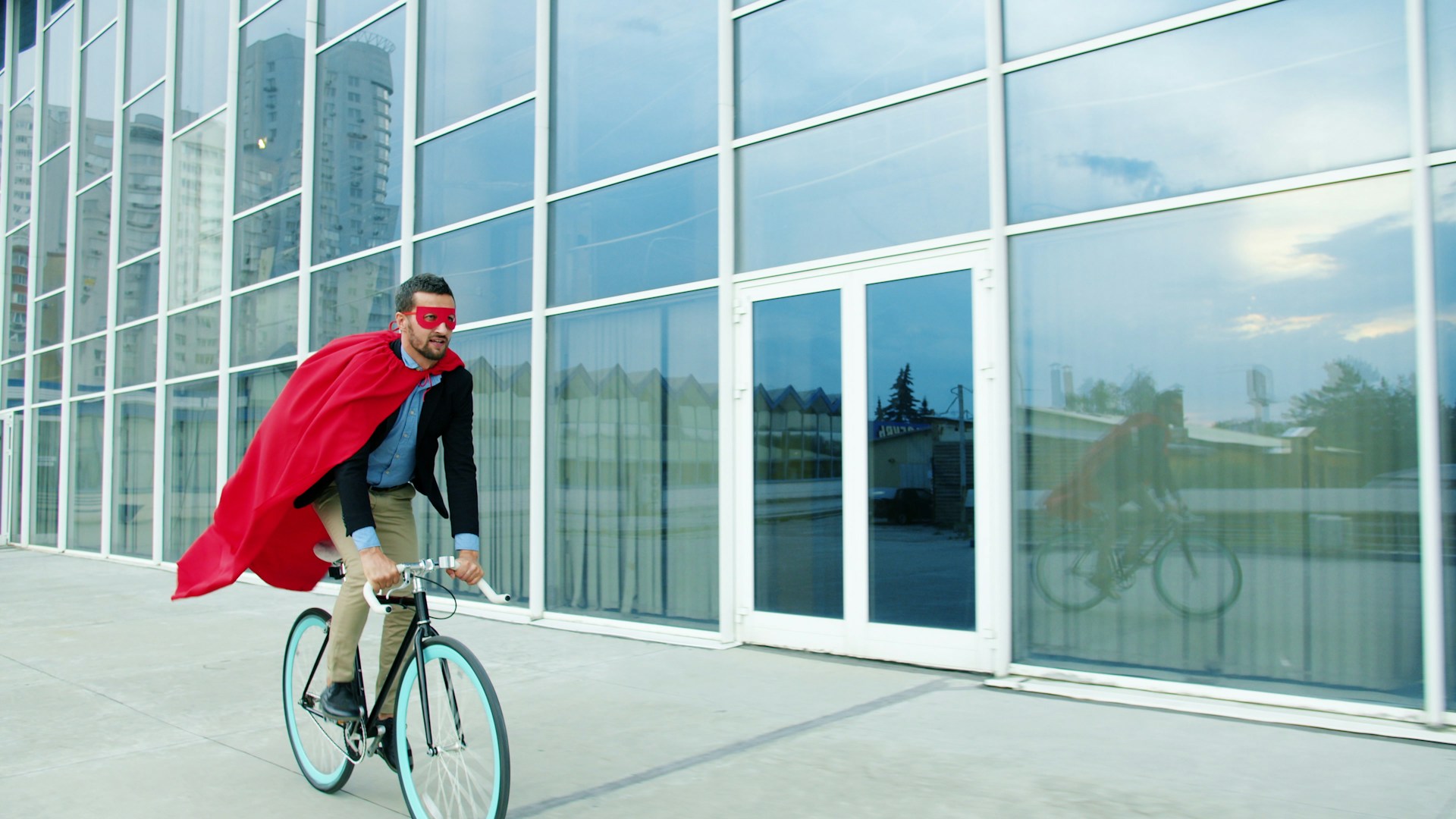Man in superhero costume rides bicycle past modern building