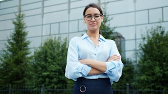 A woman in glasses stands with arms crossed outdoors.