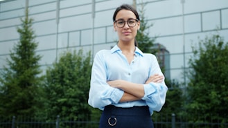 A woman in glasses stands with arms crossed outdoors.