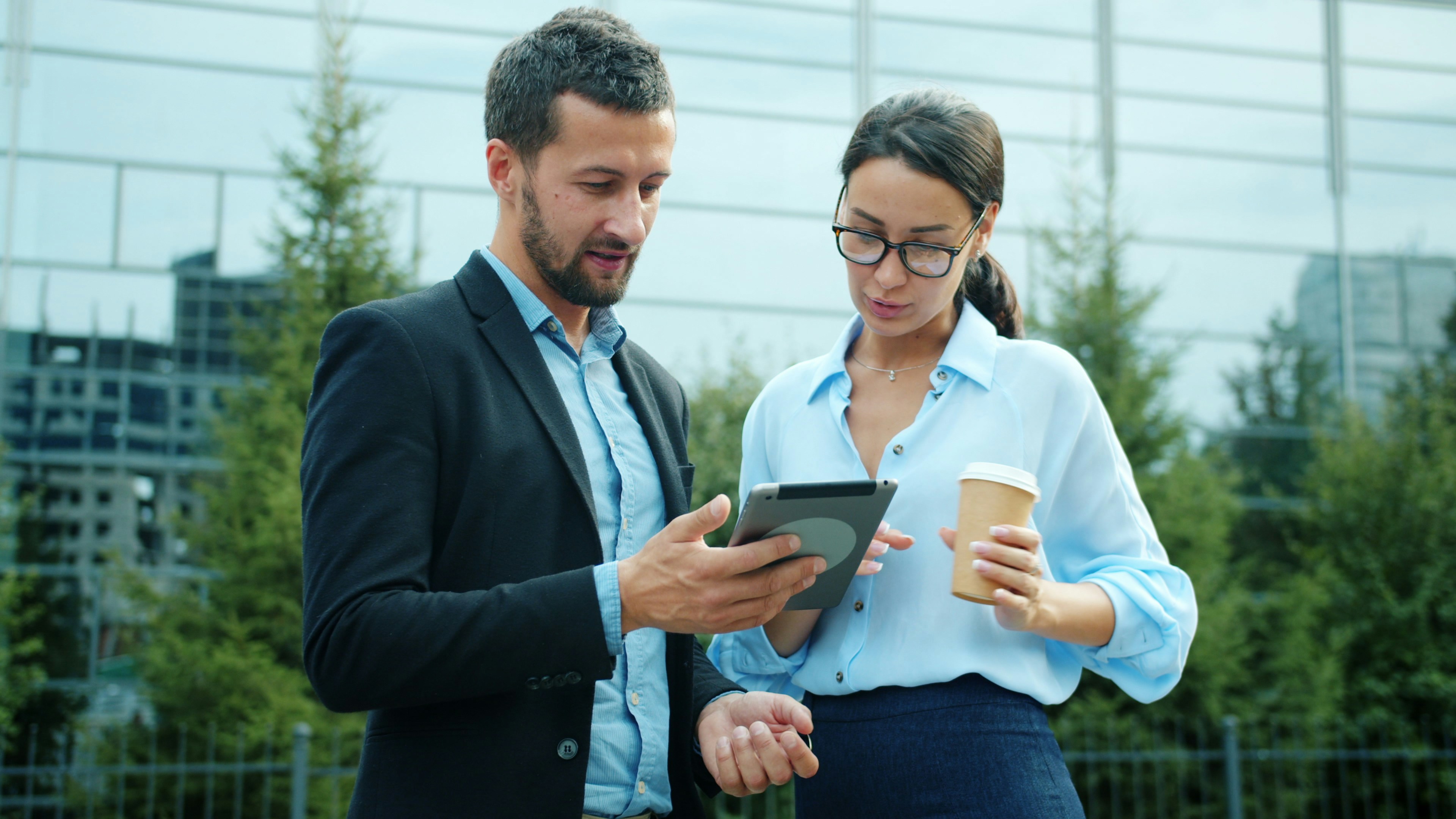 Businessman handsome young man is using tablet computer outdoors talking to cheerful woman holding take out coffee, business center is in background. People and work concept.