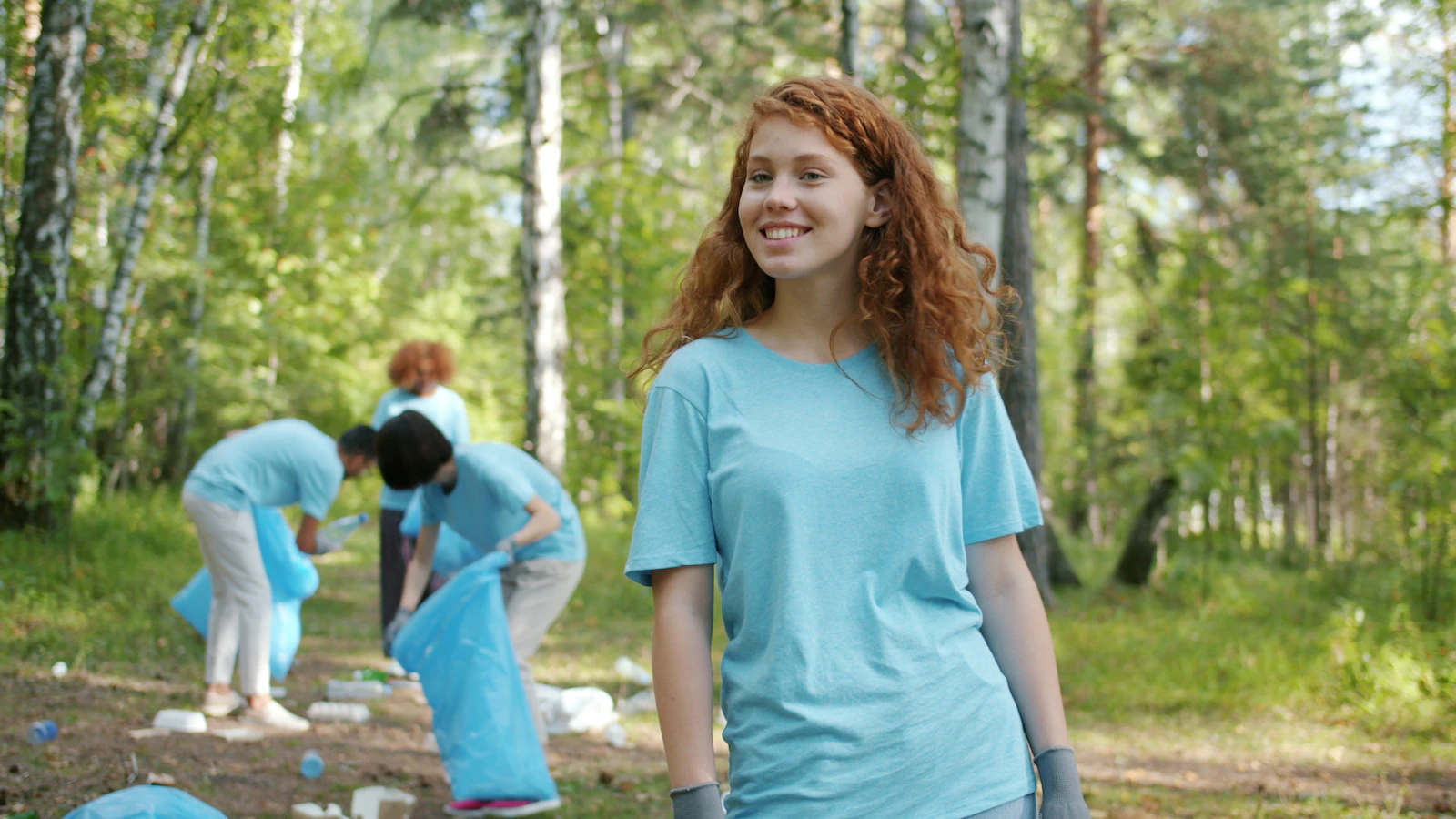 A young woman smiling while volunteering with a group cleaning up litter in a forest.