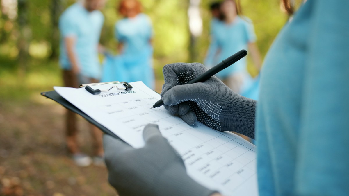 Person in gloves writing on a clipboard with a volunteer sign up sheet, volunteers working in the background