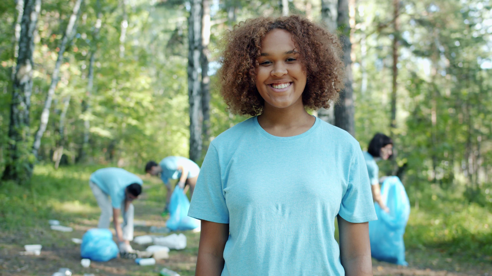 A young woman volunteer smiles at the camera while a group cleans up litter in a forest behind her.