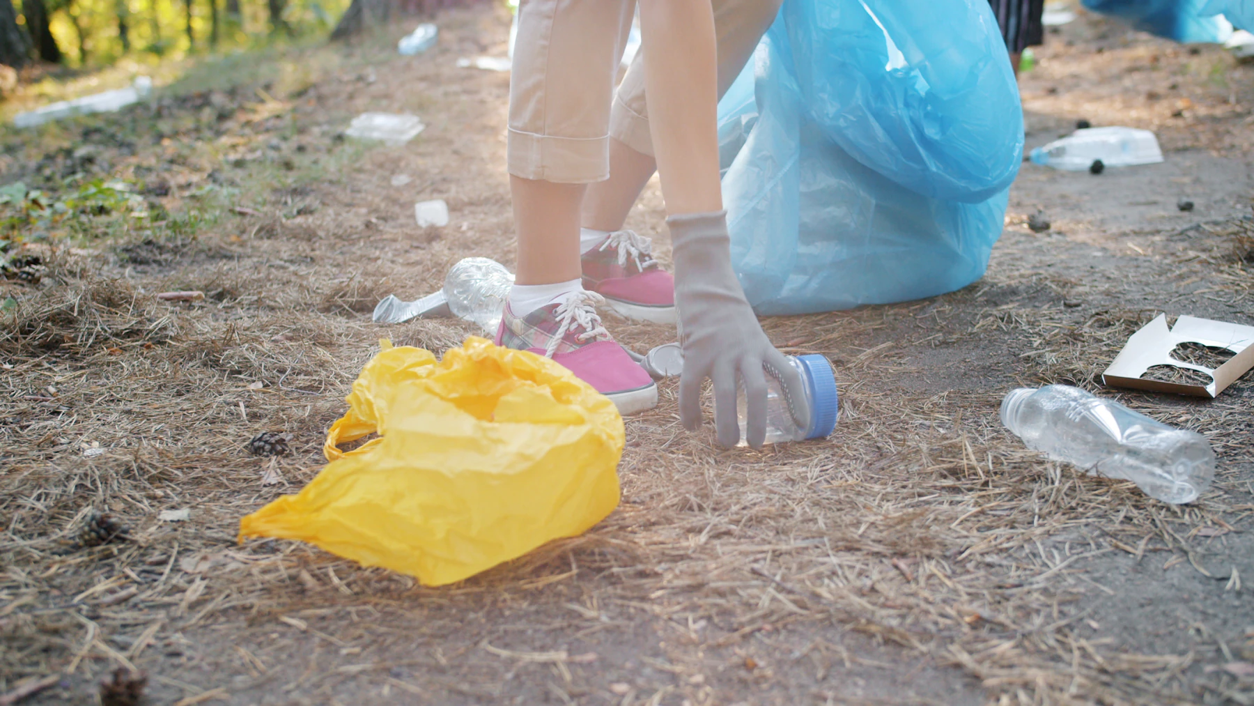 A gloved hand picking a plastic bottle off dry ground beside a blue trash bag, more litter scattered behind