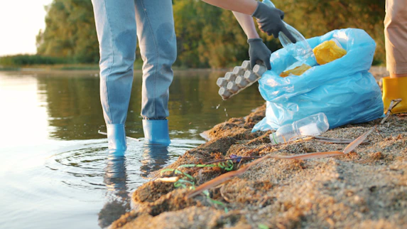 People cleaning trash from a riverbank with a blue bag.