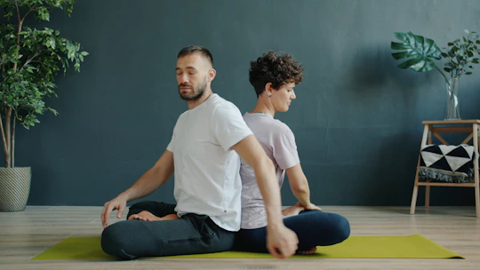 Dos personas meditando espalda con espalda sobre colchonetas de yoga.