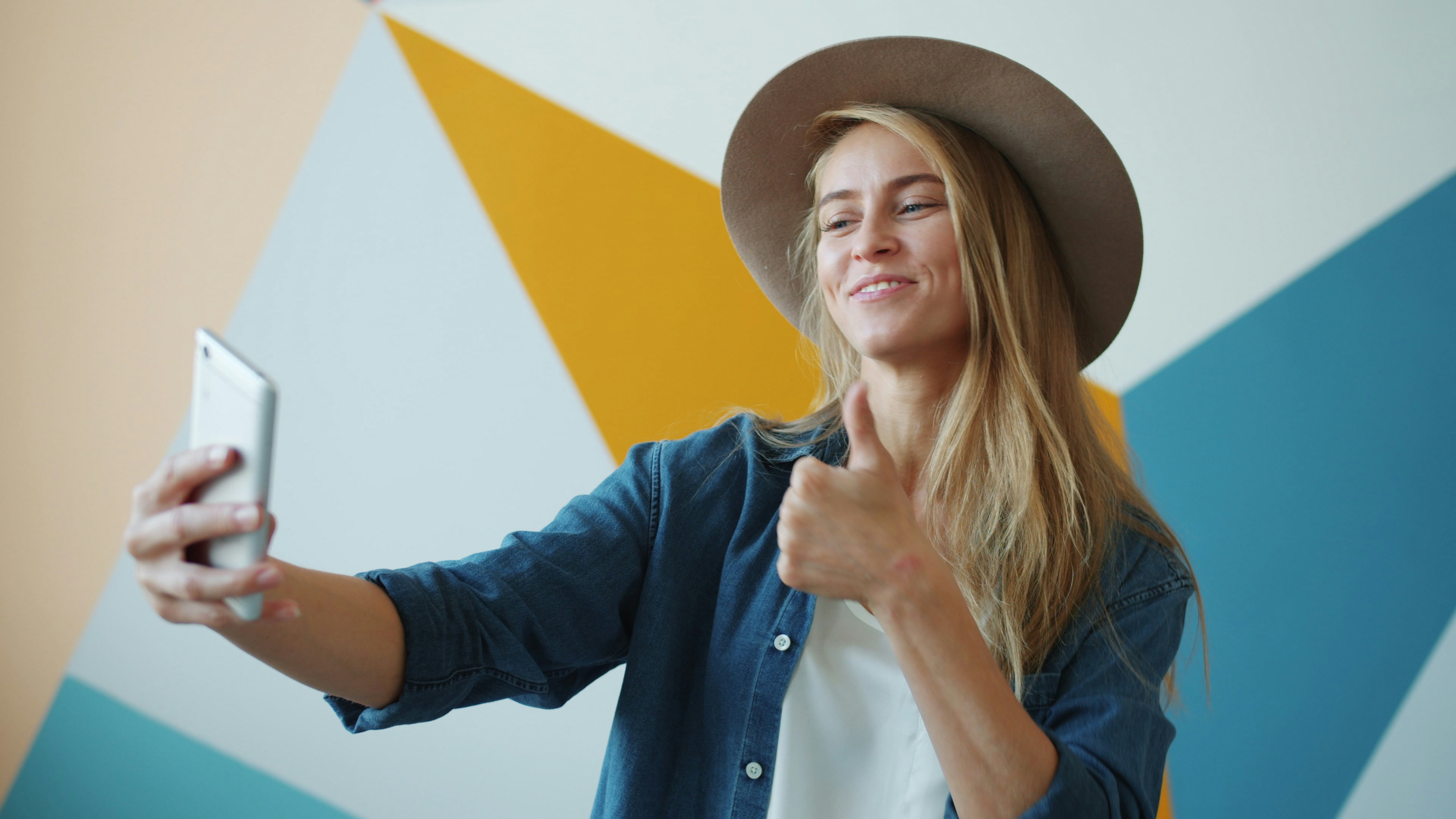 Attractive young woman wearing hat taking selfie with smartphone camera posing with hand gestures smiling showing thumbs-up. People and photograph concept.