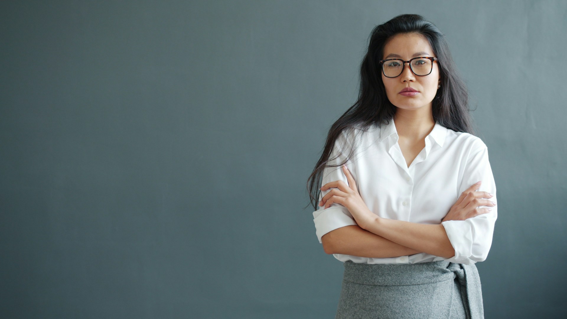 Woman with glasses and arms crossed against gray wall.