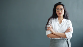 Woman with glasses and arms crossed against gray wall.