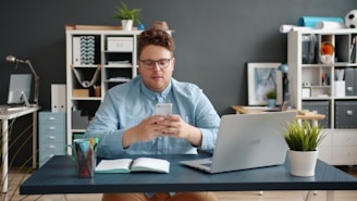 Man looking at phone at desk with laptop.