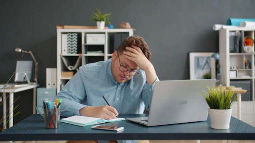 Man writing at desk with laptop, looking stressed.