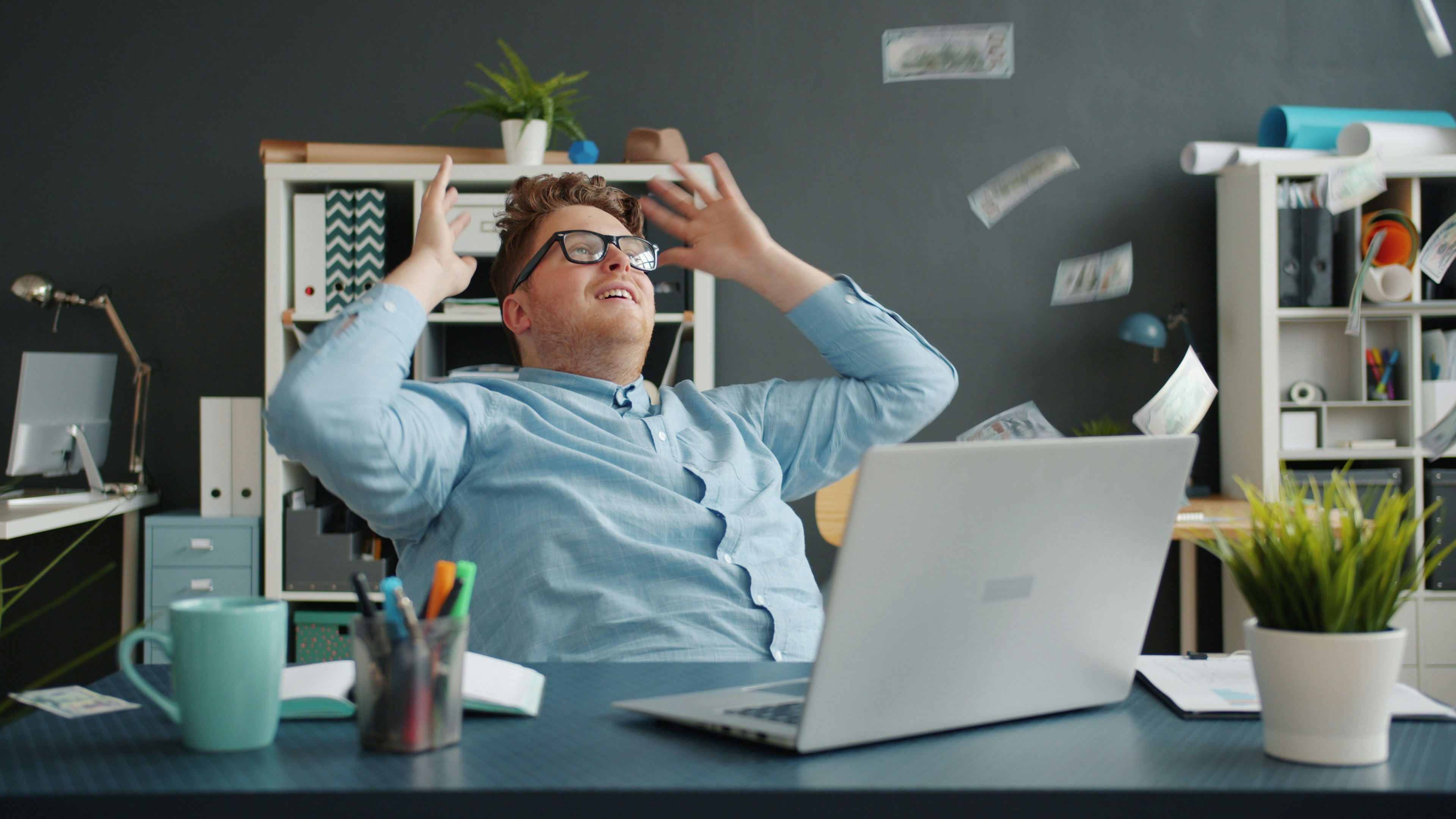 Entrepreneur attractive guy is relaxing in chair in office while money banknotes are falling down from above. Happy young people, wealth and business concept.