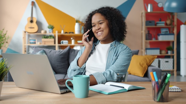 Woman talking on phone while working on laptop at desk.