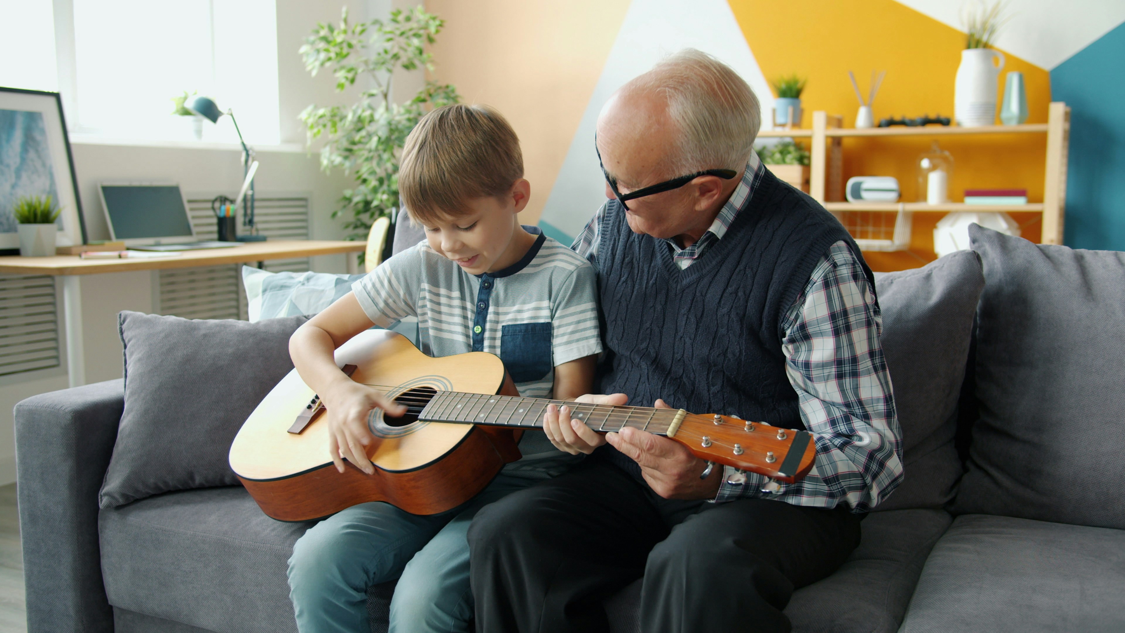 Grandfather teaching grandson to play guitar on couch.