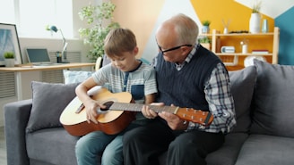 Grandfather teaching grandson to play guitar on couch.