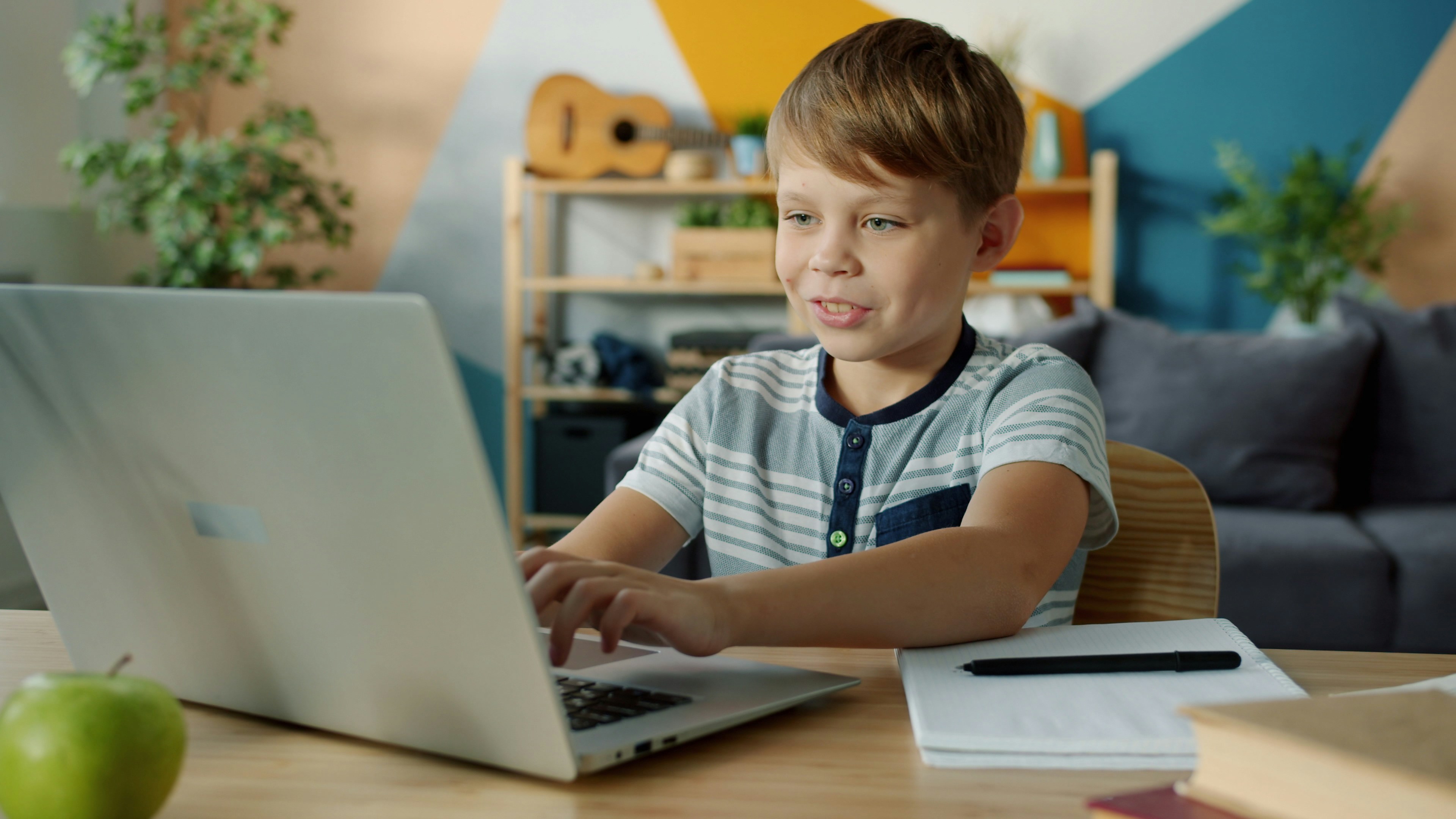Joyful child is busy doing homework using laptop typing then taking notes in notebook writing sitting at desk at home. Education and modern technology concept.