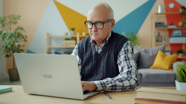 Elderly man wearing glasses using a laptop at home.