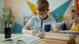 Young boy with glasses writing at desk with books.