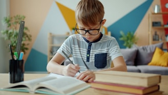 Young boy with glasses writing at desk with books.