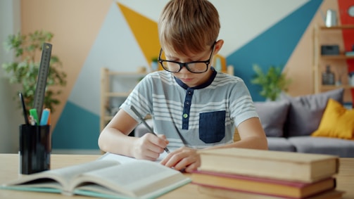 Young boy with glasses writing at desk with books.