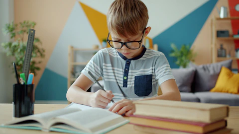 Young boy with glasses writing at desk with books