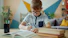 Young boy with glasses writing at desk with books.