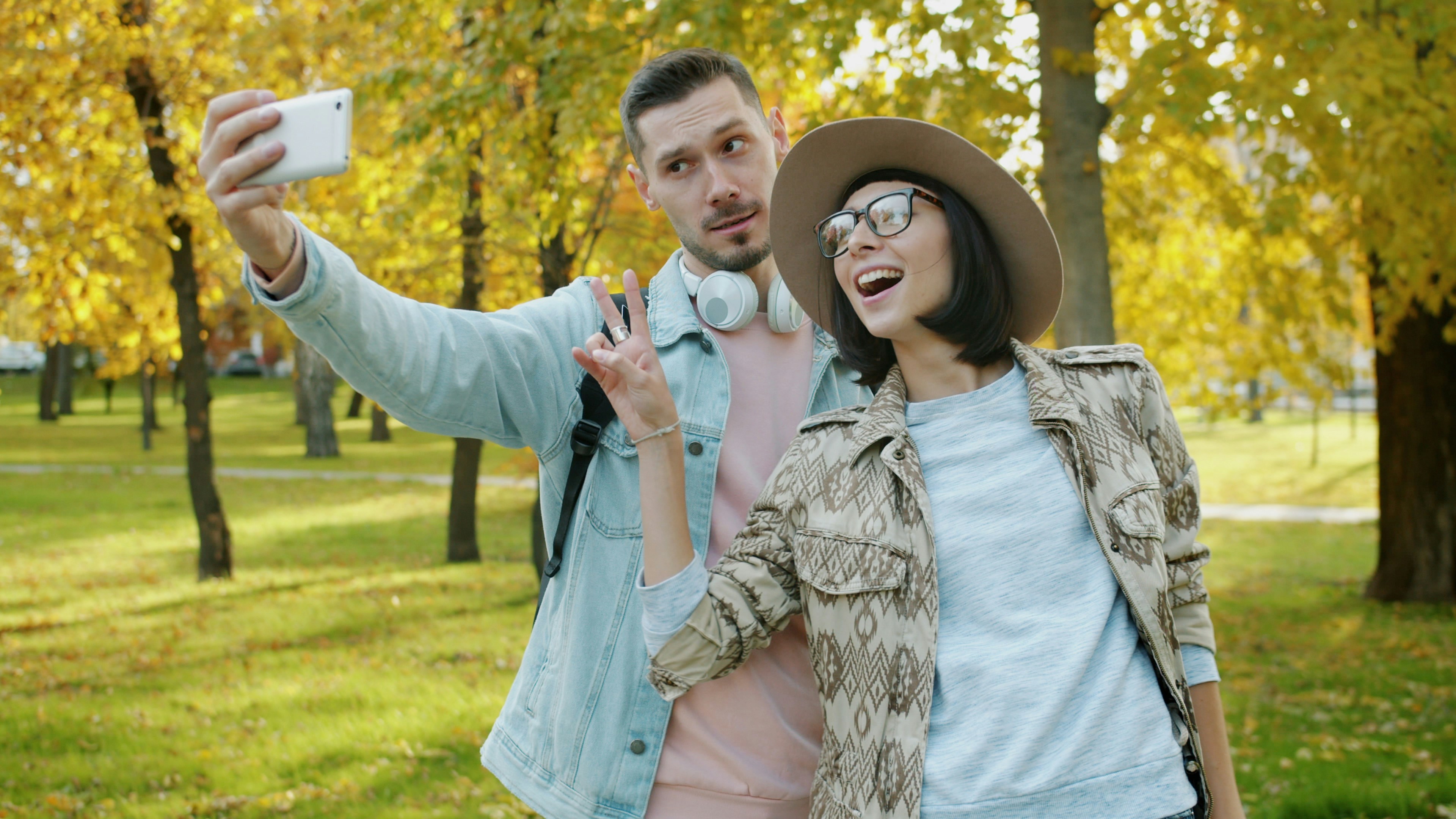 Portrait of cheerful couple taking selfie in park showing thumbs-up laughing using smartphone camera outdoors. Contemporary lifestyle and happiness concept.