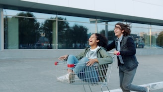 Two people riding in a shopping cart outside.