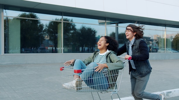 Two people riding in a shopping cart outside.