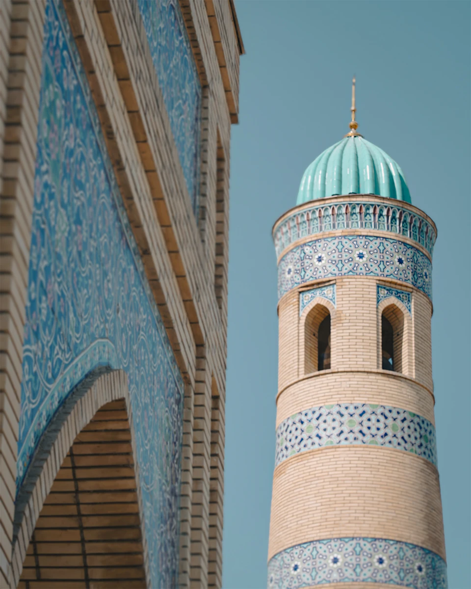 The image shows a tall, ornate brick tower with a green dome and blue tiled roof, standing against a clear blue sky. The tower is adorned with intricate patterns and designs on its facade, adding to its majestic appearance. In the foreground, there's an archway that frames the tower, creating a sense of depth in the image.

The perspective of the photo is from below, looking up at the tower, which emphasizes its height and grandeur. The colors in the image are vibrant, with the green dome contrasting against the blue sky and the blue tiled roof adding to the visual appeal.