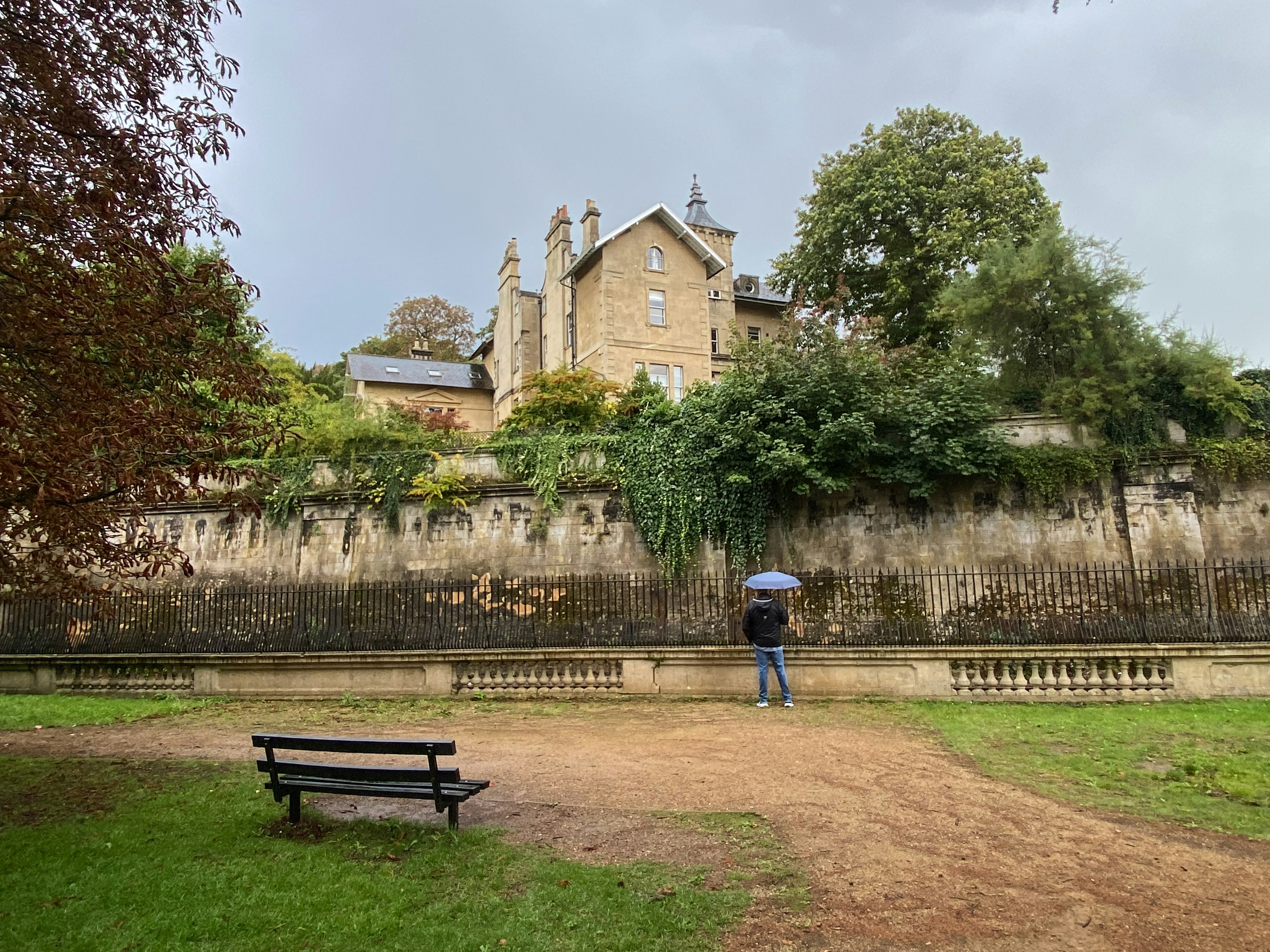 Person with umbrella near old building and park bench.