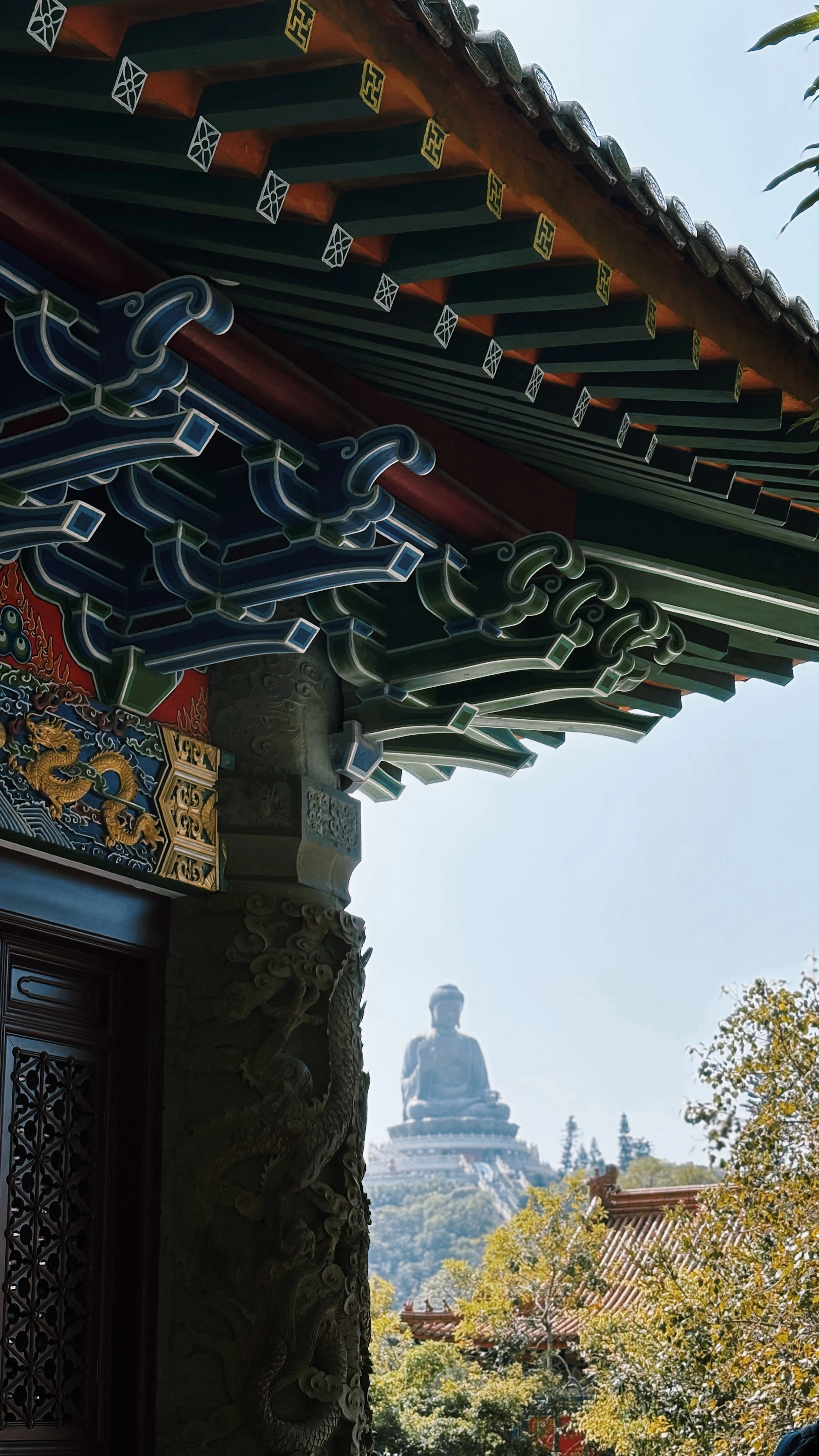 The painted green and gold eaves of Po Lin Monastery at Ngong Ping, with a carved dragon panel on the pillar below, framing the Tian Tan Big Buddha seated on its hilltop in the clear light beyond.