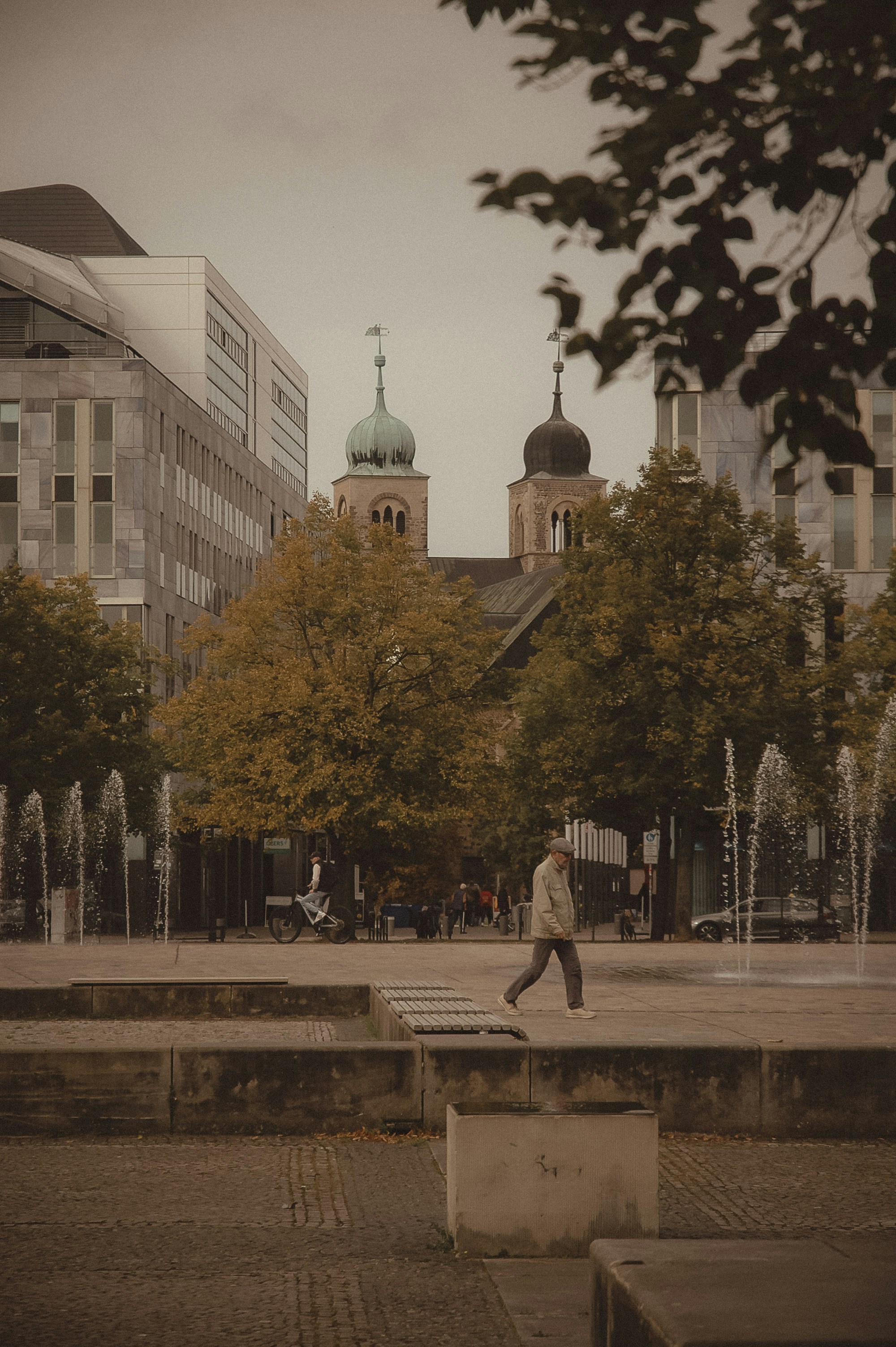 Person walking in a plaza with fountains and buildings.