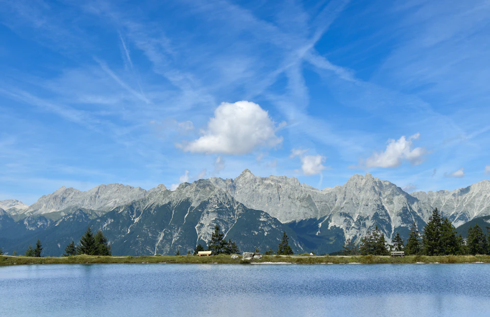 The image depicts a serene mountain landscape with a large lake at its center, surrounded by majestic mountains and clear blue skies. The mountains are covered in lush green trees, creating a beautiful contrast against the sky. A few clouds can be seen scattered across the sky, adding depth to the scene. In the foreground of the image, there is a small body of water that reflects the surrounding scenery.