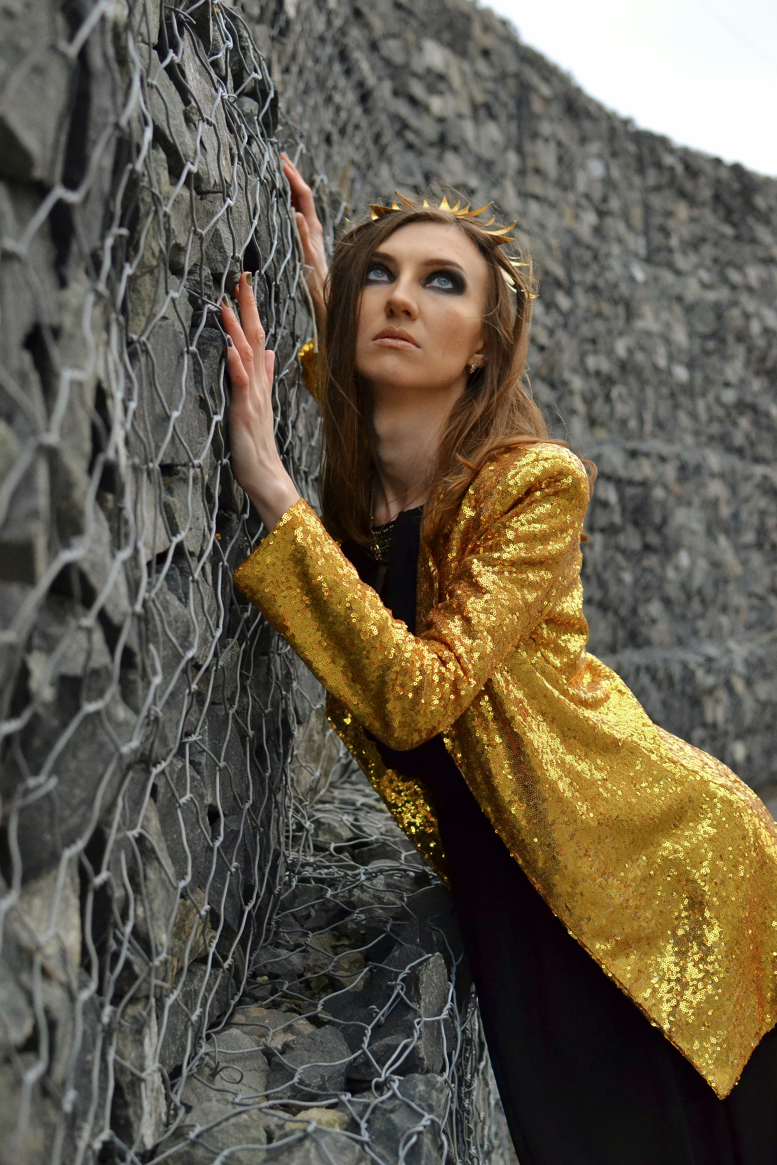 Woman with golden crown and jacket leans on wall.